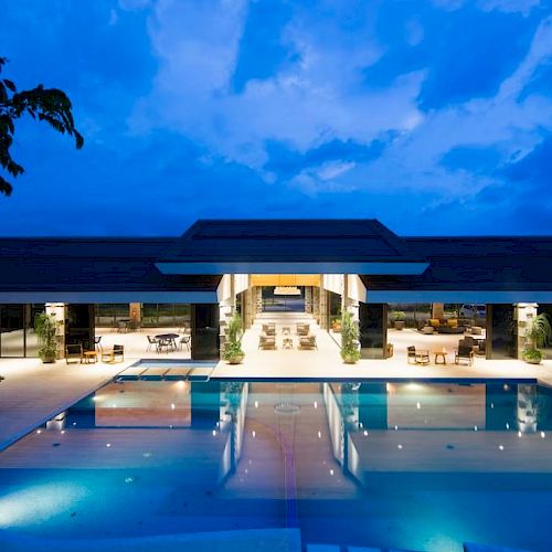 The image shows a modern building with a large illuminated swimming pool in front, set against a twilight sky.