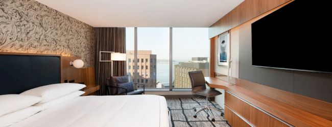 A modern hotel room with a large bed, wall-mounted TV, desk, chair, and floor-to-ceiling windows offering a city view.