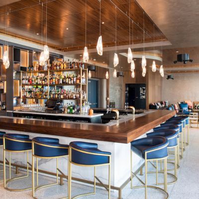 A modern bar with blue stools and a central counter, featuring hanging lights and a well-stocked bar in a bright, spacious room.