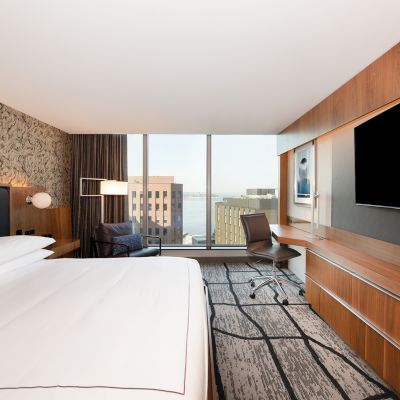 A modern hotel room with a large bed, wall-mounted TV, desk, and chair, offering a view of buildings through a large window.