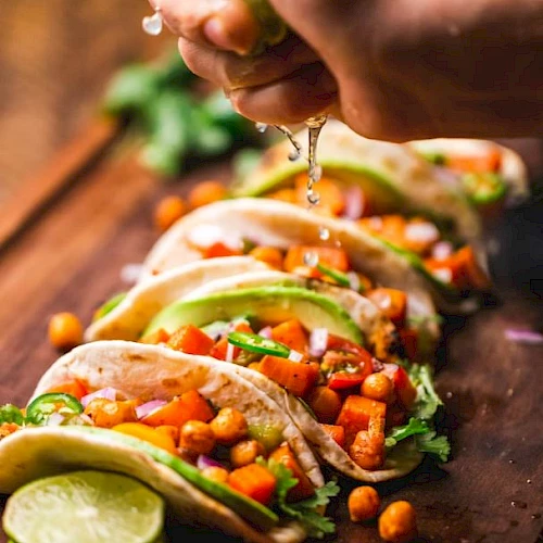 A hand squeezes lime over tacos filled with chickpeas, avocado, and vegetables on a wooden board.