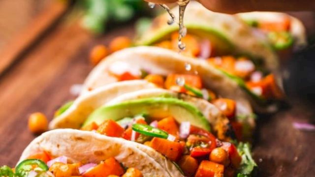 A hand squeezes lime over tacos filled with chickpeas, avocado, and vegetables on a wooden board.