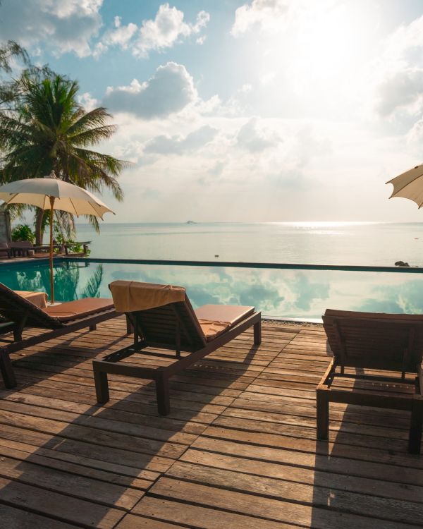 Lounge chairs and umbrellas by a pool overlook a tranquil sea, framed by palm trees and a bright, partly cloudy sky.