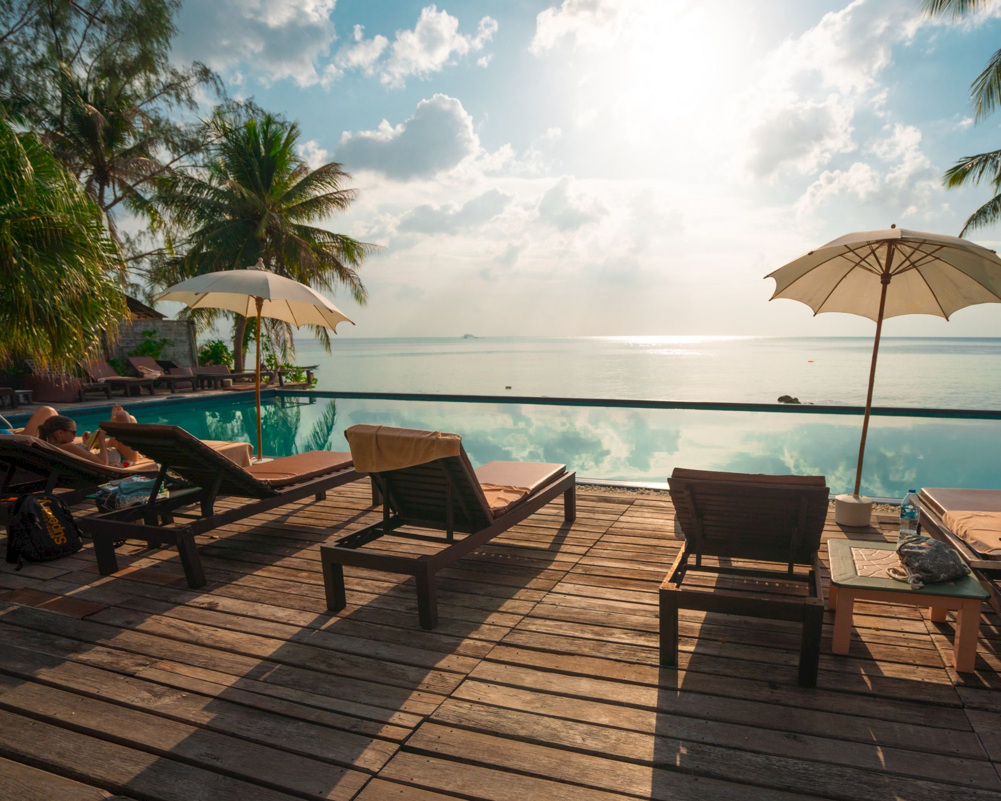 Poolside scene with lounge chairs, umbrellas, overlooking a calm ocean and palm trees under a partly cloudy sky.