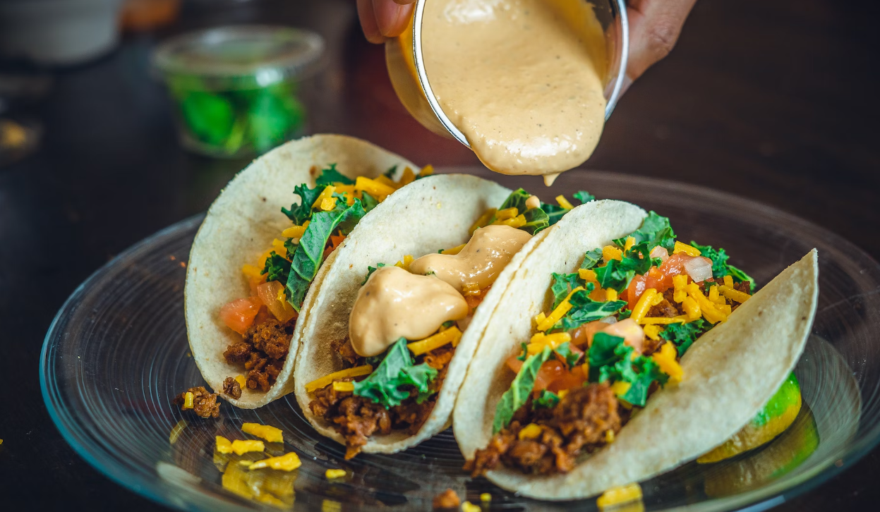 Three tacos filled with meat, cheese, and lettuce, with sauce being poured over them on a plate.
