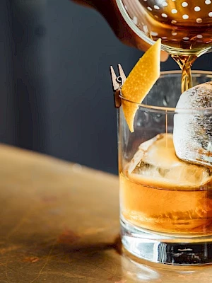A cocktail being poured into a glass with ice and a twist of citrus peel, sitting on a bar countertop.