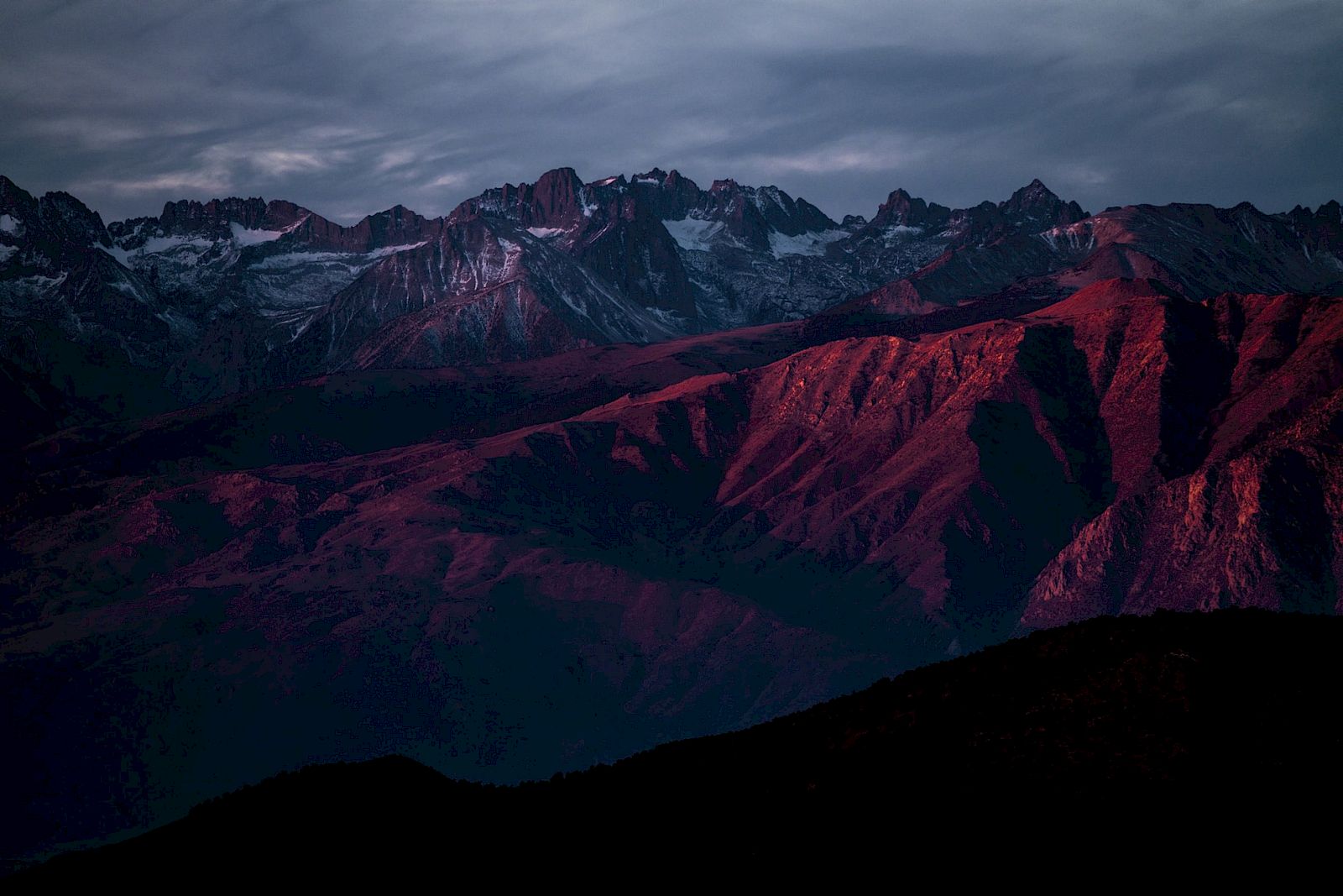 A mountain range at dusk or dawn with deep red and purple hues, under a cloudy sky.