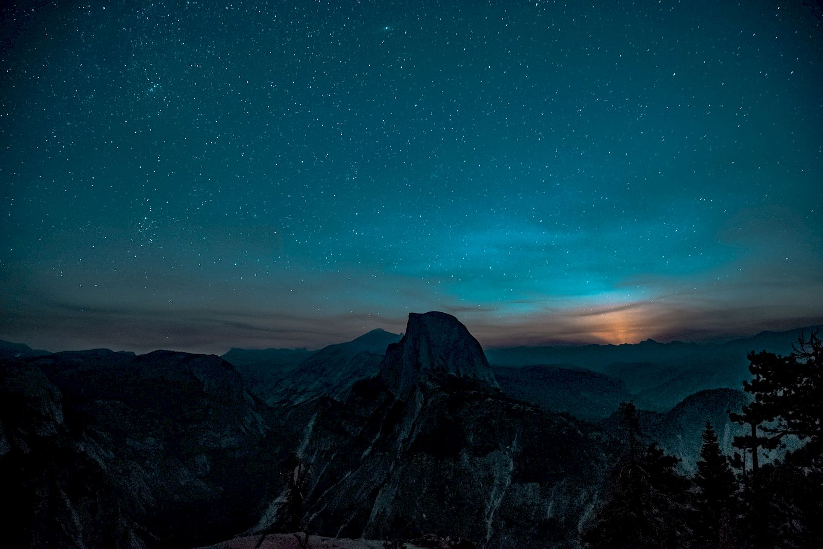A night sky filled with stars over a mountainous landscape with a glowing horizon, possibly indicating a sunset or sunrise.