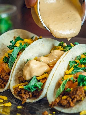 Three tacos with meat, cheese, and greens are on a plate, with sauce being poured over them from a small container.
