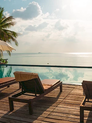 A tranquil poolside scene by the ocean with sun loungers, umbrellas, and palm trees, under a partly cloudy sky.