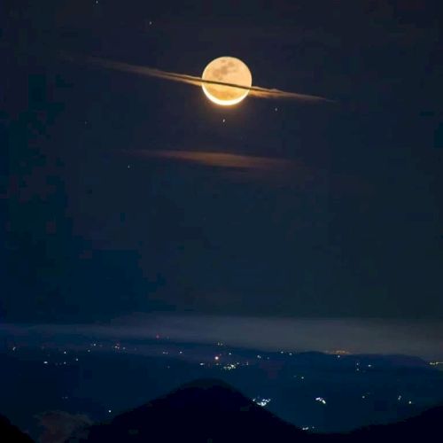 A full moon appears surrounded by clouds, resembling Saturn, with hills and city lights in the foreground.