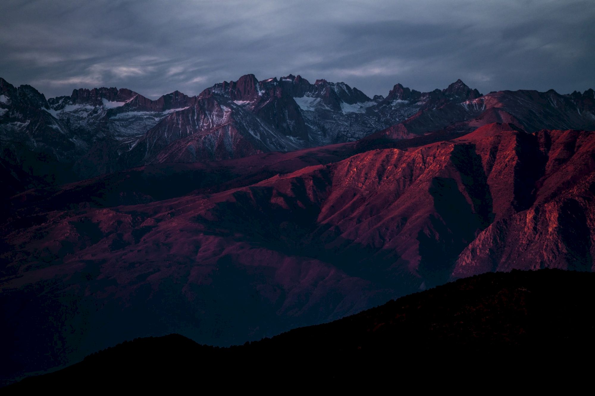 A mountain range at sunset with red and purple hues under a cloudy sky.