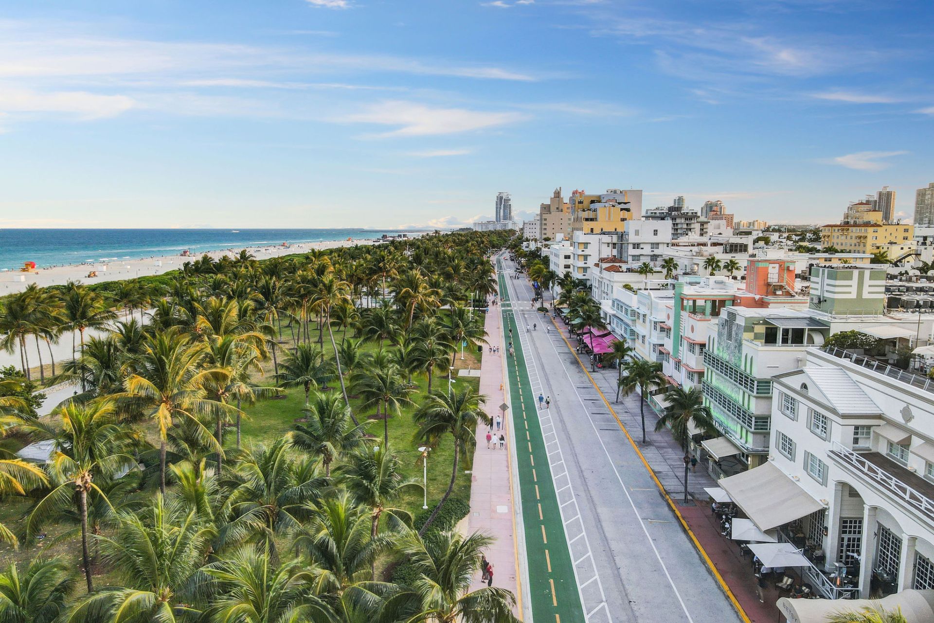 A coastal city scene with a beach, palm trees, a road with a bike lane, and buildings lining the street.