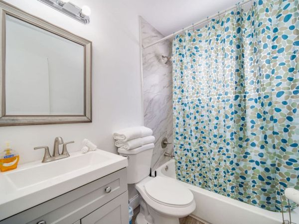 A clean bathroom with a sink, mirror, toilet, and a bath with a blue-green leaf-patterned shower curtain, plus folded towels and soap.