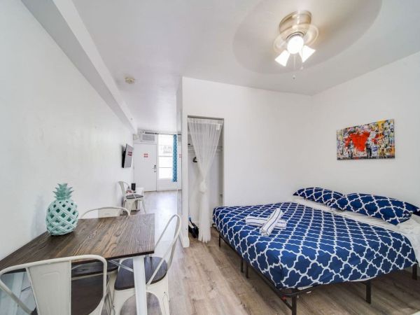 The image shows a bedroom with a bed, patterned blue bedding, a table, chairs, wall art, and a ceiling fan.