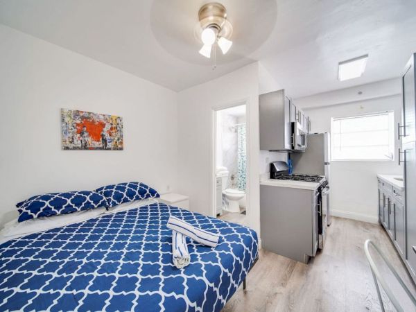 A small apartment with a bed, artwork, bathroom, and kitchen, featuring blue bedding, white walls, and modern appliances.