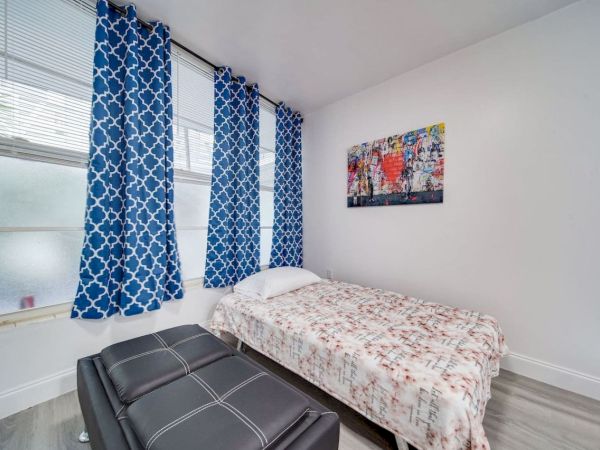 A small bedroom with blue curtains, single bed, patterned bedding, a wall art piece, and a black ottoman on light-colored flooring.