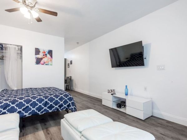 A bedroom with a blue-patterned bed, wall-mounted TV, dresser, and wall art. A fan and a LOVE decor item are visible.