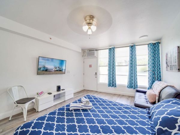 A bright room features a bed, a sofa, a wall-mounted TV, and a small table with a chair, complete with patterned curtains and decor.