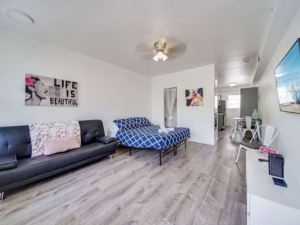 The image shows a modern room with a sofa, bed, wall art, and a view of a kitchen area, all in a minimalist style.