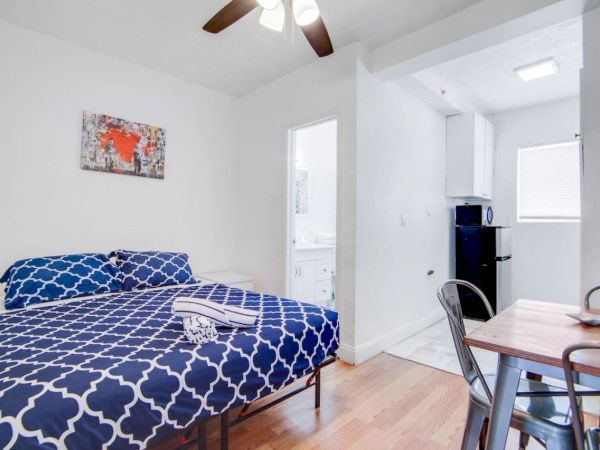 The image shows a small bedroom with a blue bedspread, a table and chairs, a bathroom door, and a kitchenette with a fridge.