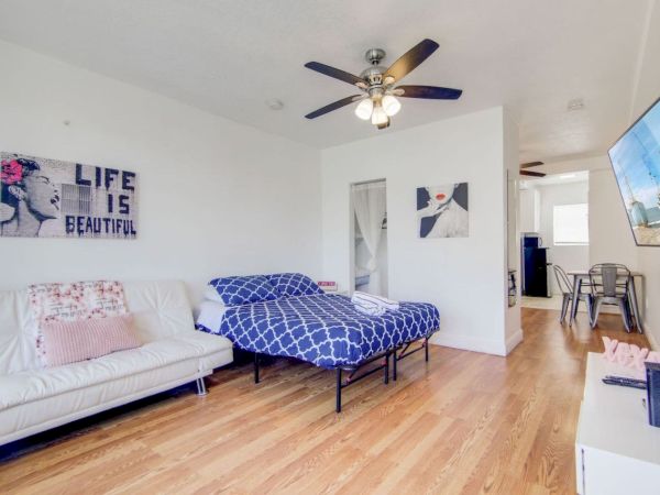 A living area with a sofa, bed, wall art, TV, and ceiling fan. Light wooden floor and dining table in the background complete the space.