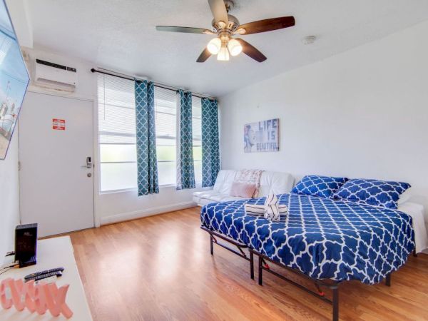 A room with a bed, blue patterned bedding, wall art, TV, ceiling fan, sofa, and large windows on a wooden floor.