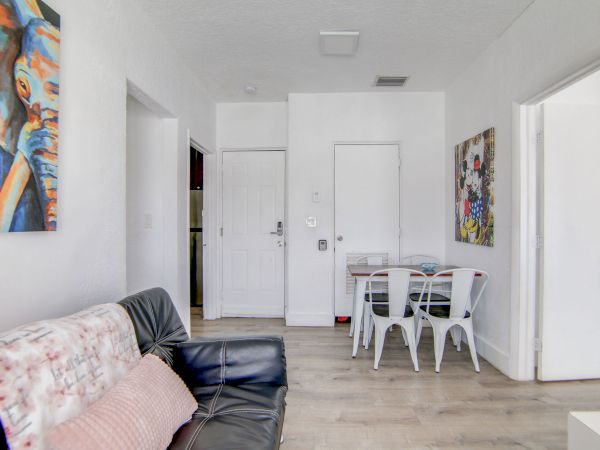 A living room with a black sofa, wall art, a small dining area with four chairs, and white walls and wooden flooring.