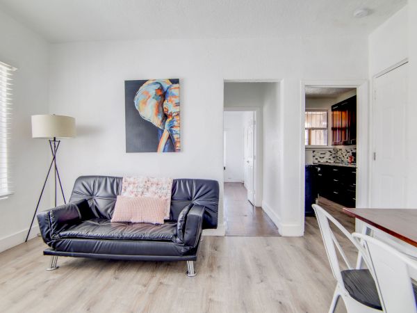 A modern living room with a black sofa, colorful artwork, a floor lamp, and light wooden flooring. A kitchen is visible through a doorway.