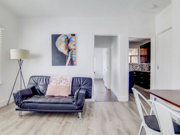 A modern living space with a black sofa, pillow, floor lamp, artwork, dining table, chairs, and view towards a kitchen in the background.