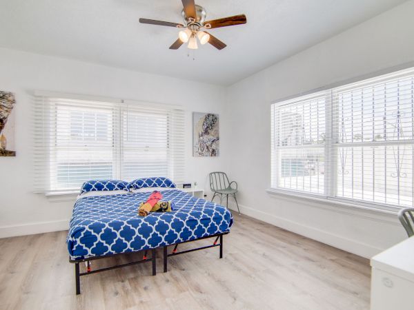 A bright bedroom with a blue-patterned bedspread, ceiling fan, large windows, and modern decor.