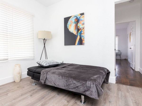 A minimalist bedroom with a low bed, elephant artwork, lamp, and blinds. A hallway leads out the room.