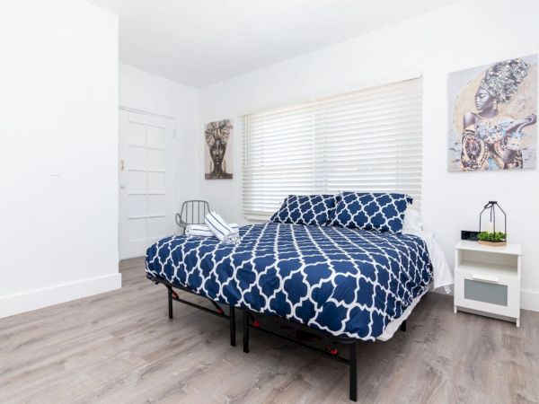 A bedroom with a bed featuring blue patterned bedding, a side table, artwork on walls, and a window with blinds.