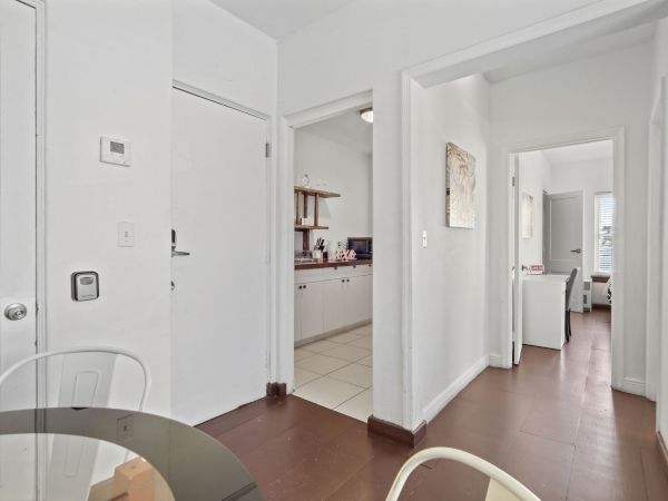 The image shows a bright, minimalistic interior with white walls, a wooden floor, and doors leading to other rooms.
