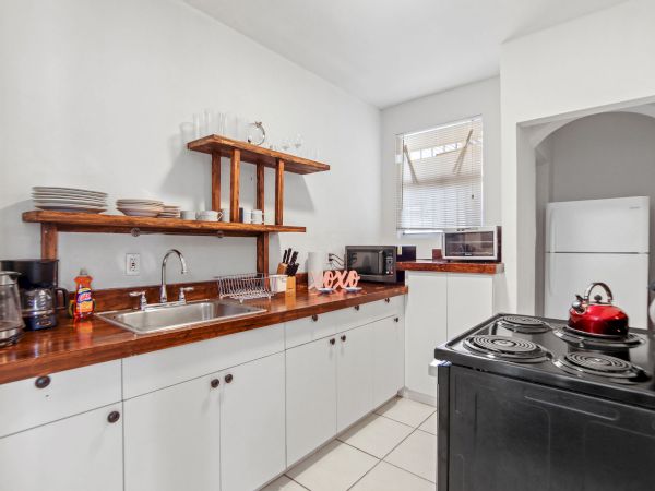 A kitchen with white cabinets, wooden shelves, a sink, stove, and a red kettle; a window and a separate laundry area are visible.
