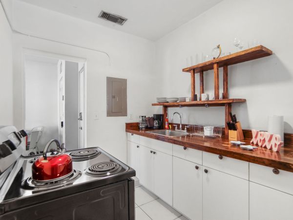 A small kitchen with white cabinets, wooden countertops, open shelves, a stove with a red pot, and various kitchen items on display.