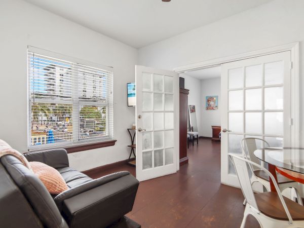 The image shows a bright living area with a sofa, glass table, chairs, and French doors leading to another room with wooden floors.