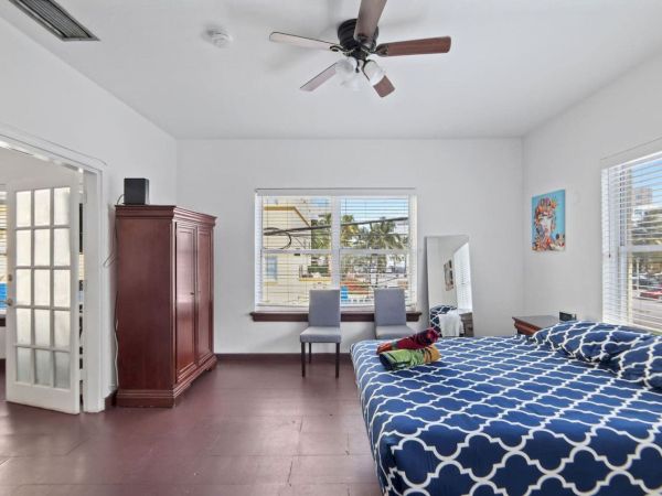 A bedroom with a blue-patterned bedspread, wooden wardrobe, ceiling fan, and windows overlooking a street view.