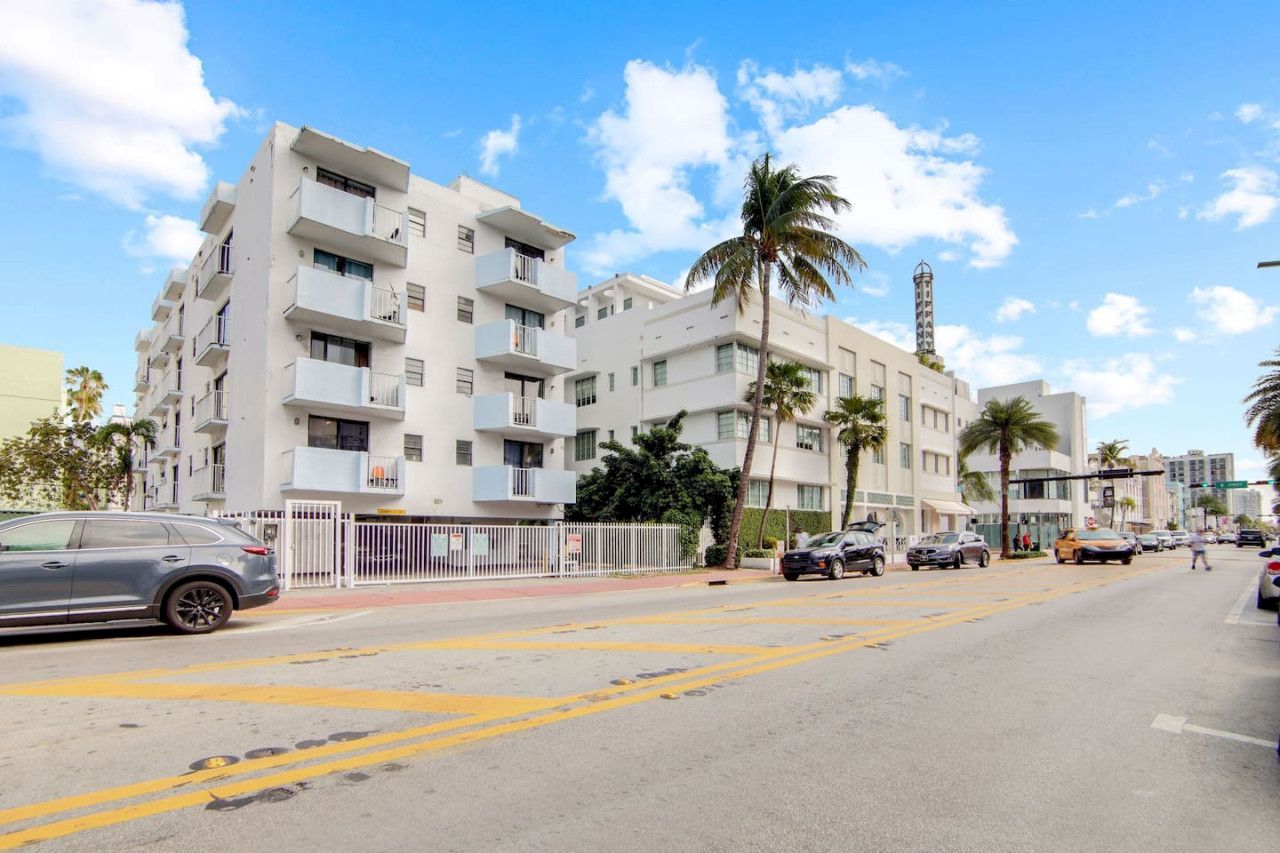 The image shows a street with mid-rise buildings, palm trees, and cars under a clear blue sky with clouds.