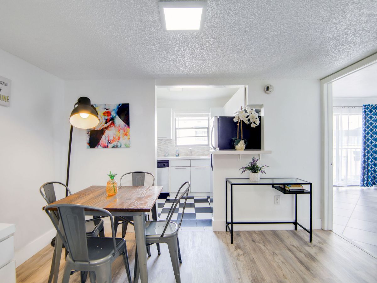 A modern dining area with a wooden table, metal chairs, a large lamp, open kitchen, artwork, and minimal decor on light wood flooring.