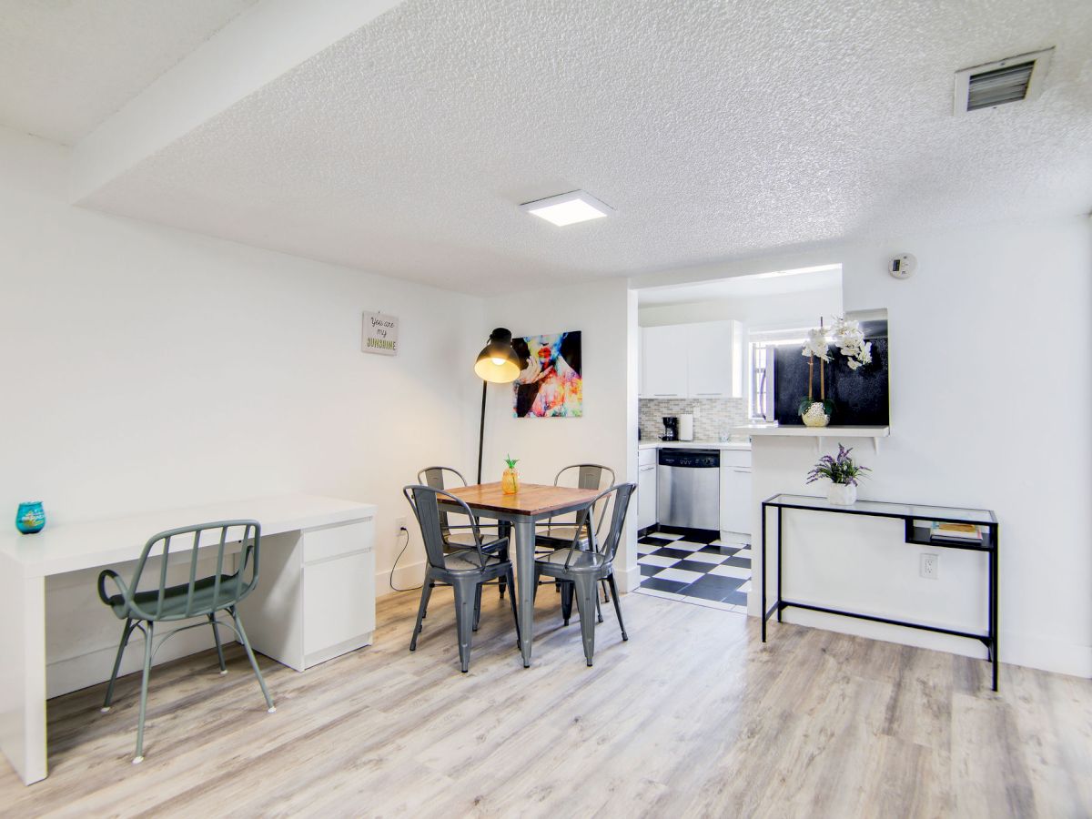 A modern dining area with a table, chairs, and a desk. The space has white walls, wooden flooring, and a view of the kitchen.