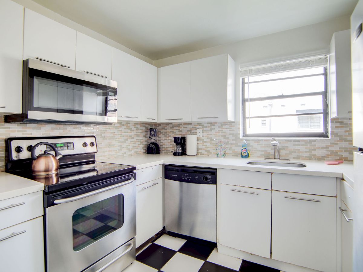 The image shows a modern kitchen with white cabinets, stainless steel appliances, a checkered floor, and a window above the sink.