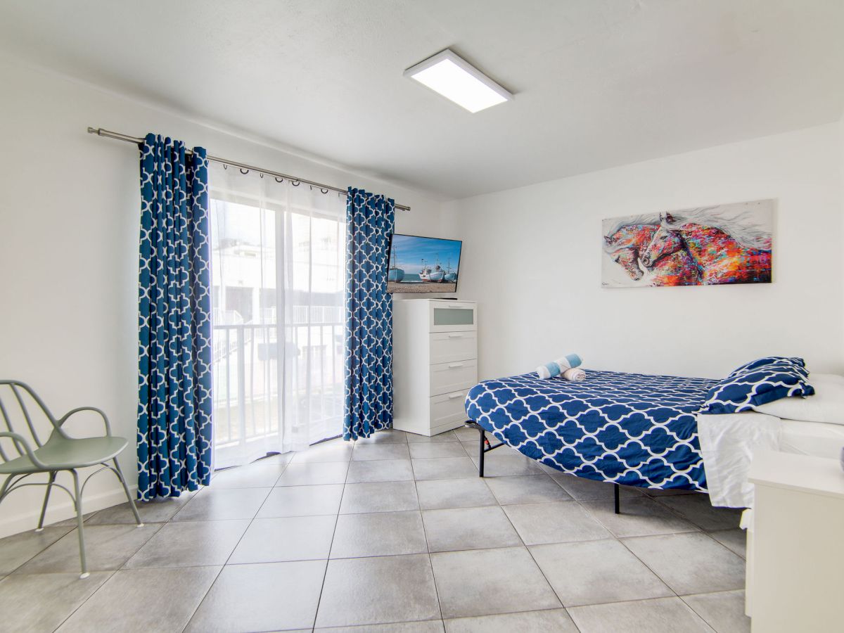 A tidy room with a bed, blue curtains, a chair, and a TV. The walls are white, and there's a window letting in natural light.