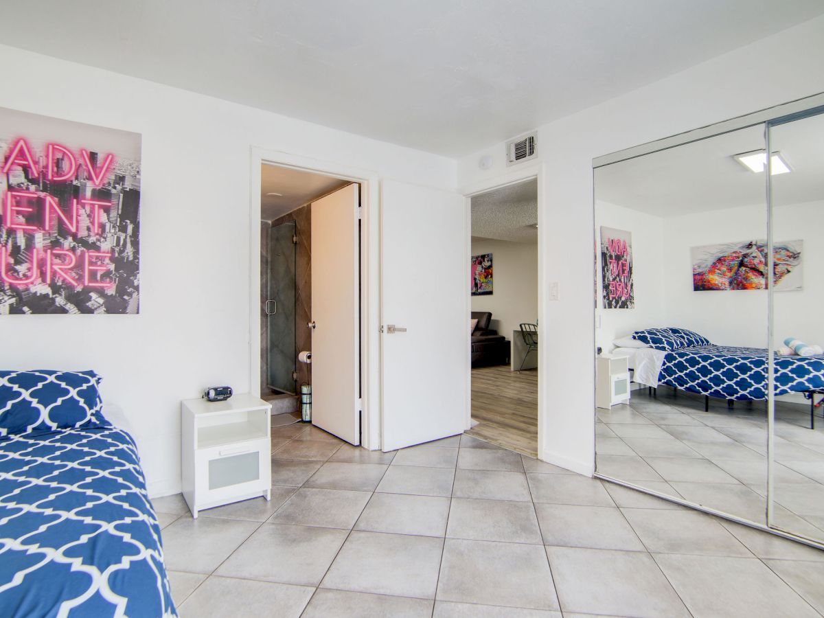 A room with a bed, tiled floor, mirrored closet, wall art, and an open door leading to another space.