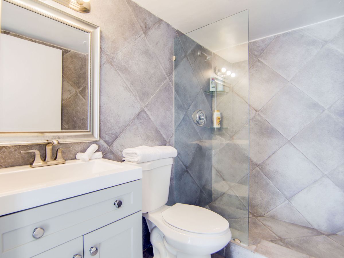 This image shows a modern bathroom with a sink, a mirror, a toilet, and a glass-enclosed shower, featuring gray tiled walls.