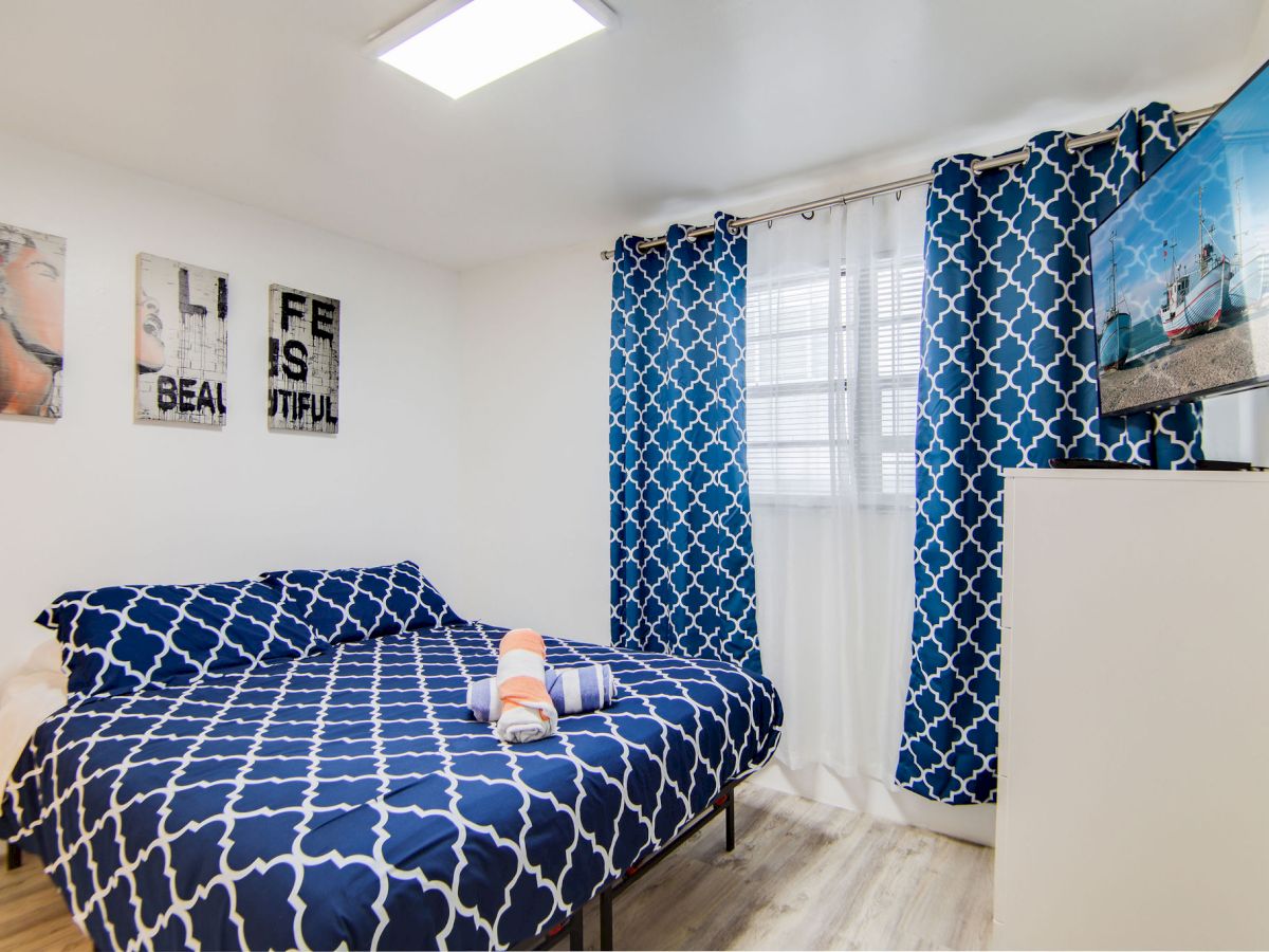 A bedroom with blue patterned bedding and curtains, wall art, a TV on a dresser, and a bright window, giving a cozy and modern vibe.