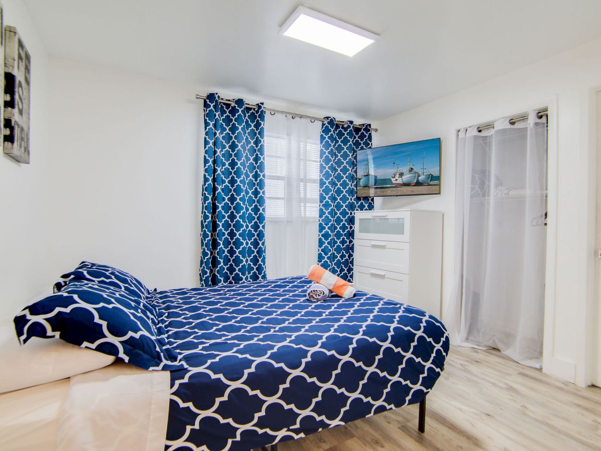 A bedroom with a blue patterned bedspread, matching curtains, dresser, and wall art. A stuffed animal and hanging wardrobe visible.
