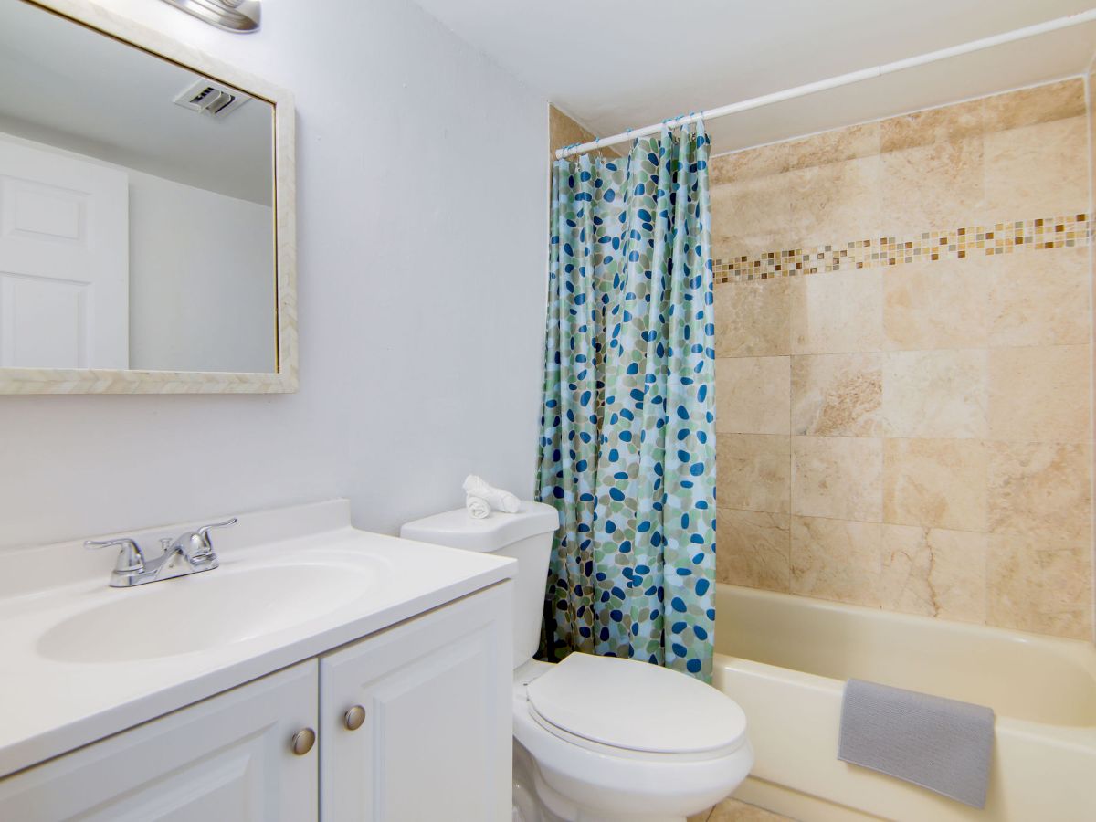 A bathroom with a sink, mirror, toilet, and a bathtub with a blue shower curtain and a gray towel hanging over the tub edge.