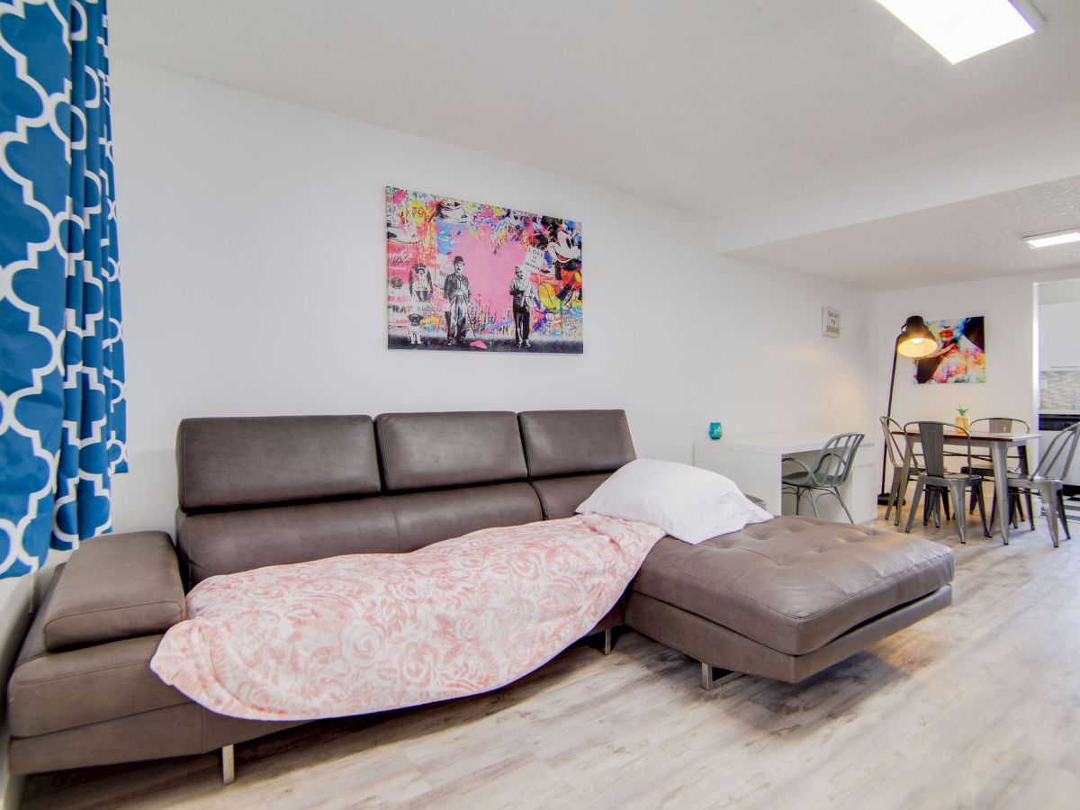 A modern living room features a gray sectional sofa, artwork, a dining area with a table and chairs, and a kitchen in the background.