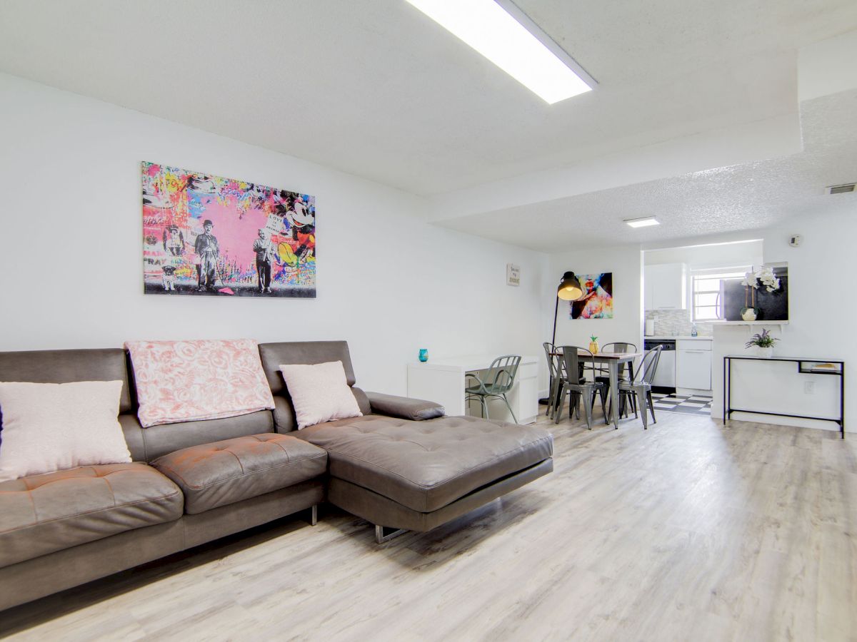 The image shows a modern living room with a brown sectional sofa, colorful wall art, a dining table, and a wall-mounted TV.